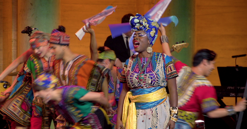 Woman sings in African costume as dancers move behind her